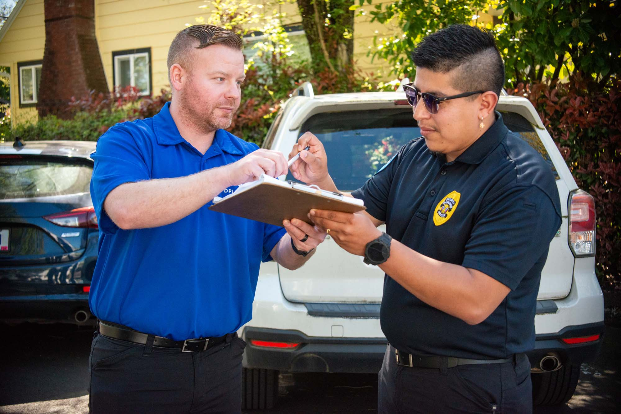 A DPI Security supervisor in a blue company polo and a uniformed security officer review paperwork on a clipboard together in a parking lot.