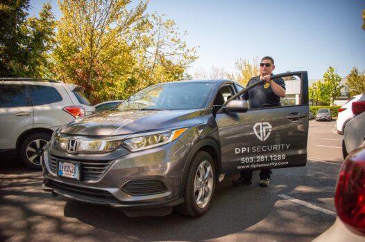 A uniformed DPI Security officer stands at the open driver’s door of a gray DPI Security patrol vehicle in a sunny commercial parking lot.